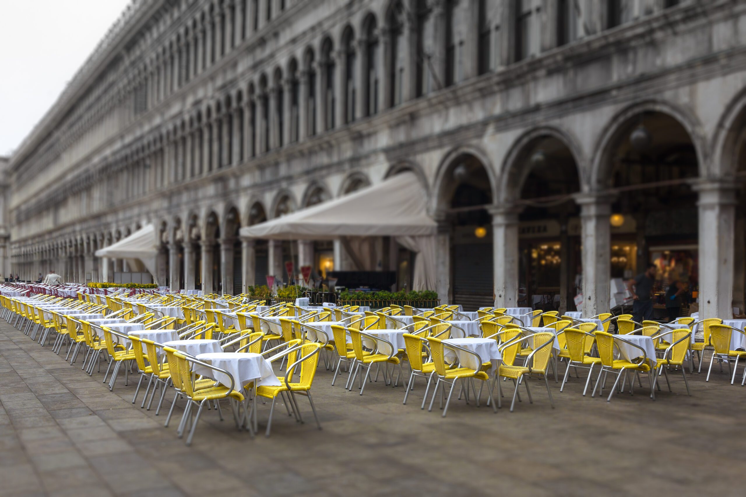 This vibrant piece captures a contrast of spirits, where radiant yellow chairs defy the grey atmosphere at Piazza San Marco,