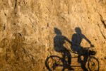 Tranquil synergy of light and shadow, as cyclists cast elongated figures on a sunlit rock wall, epitomizing Vancouver’s vibrant seawall in Stanley Park.