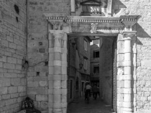 A grayscale image capturing the essence of historical Trogir, Croatia, focusing on an architectural doorway, with a local resident immersed in a moment of solitude, complemented by the cobbled street and distant human elements, symbolizing the blend of everyday life with the town's ancient allure.