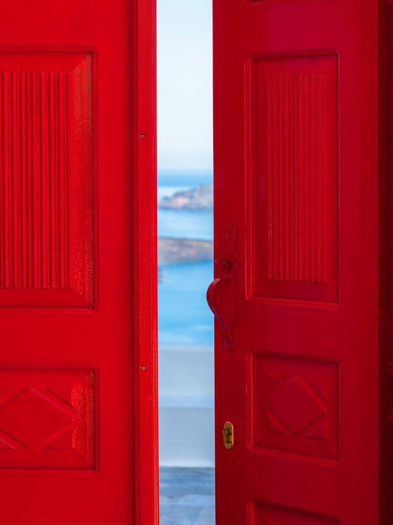 Bright red doorway opening to a view of the Santorini caldera with blue waters below.