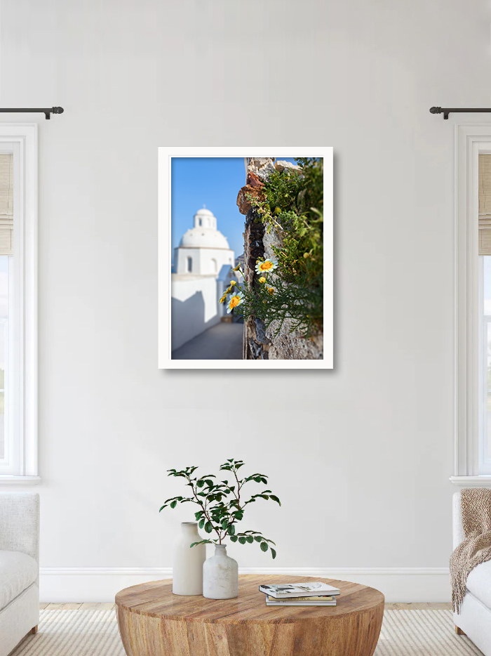 Ancient walkway in Fira, Santorini with yellow flowers and a white domed church in the distance.