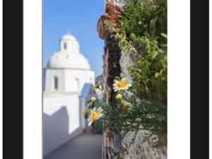 Ancient walkway in Fira, Santorini with yellow flowers and a white domed church in the distance.