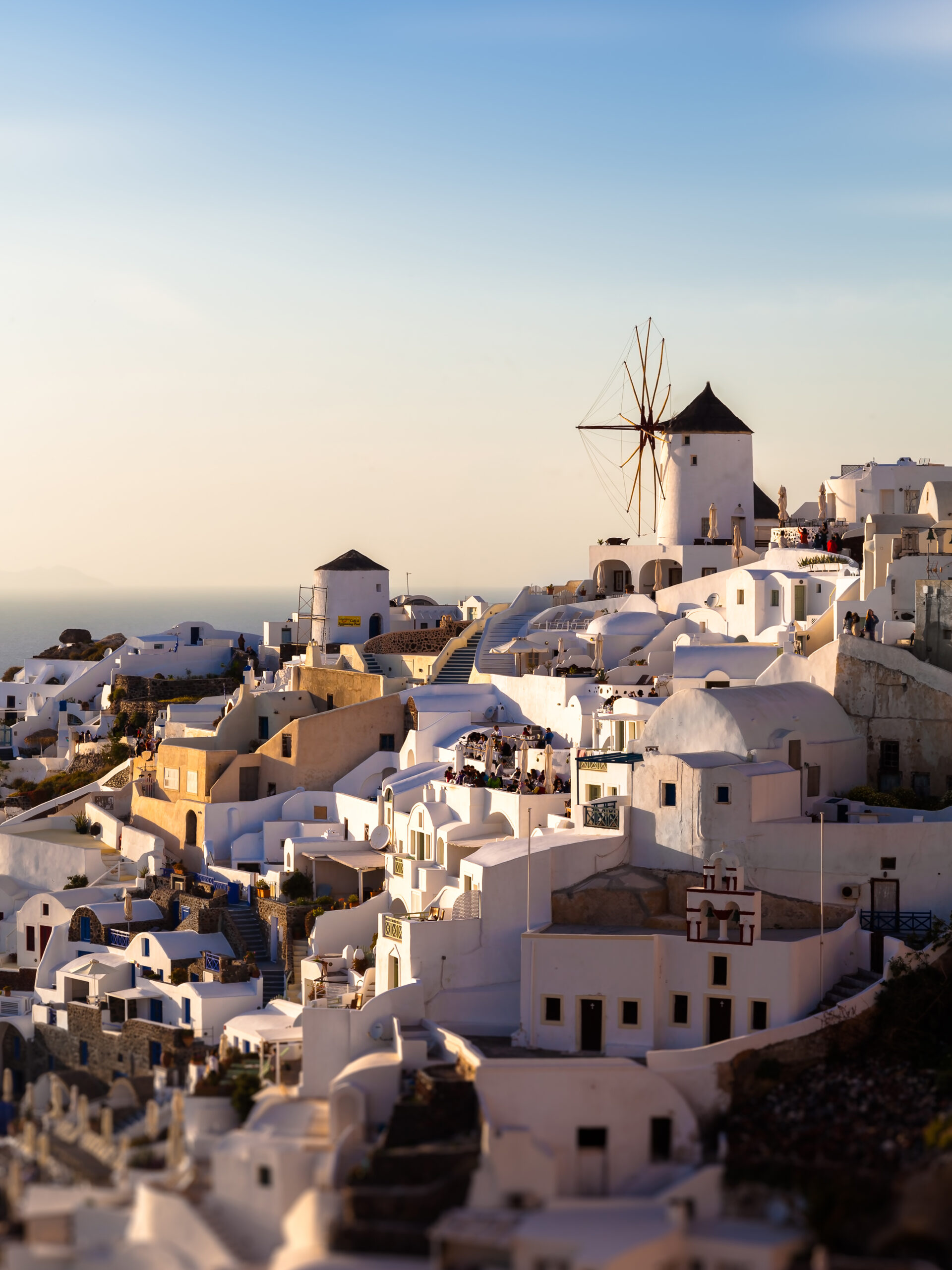 The village of Oia in Santorini, Greece, captured at dusk with soft golden lighting illuminating the iconic windmills and white buildings.