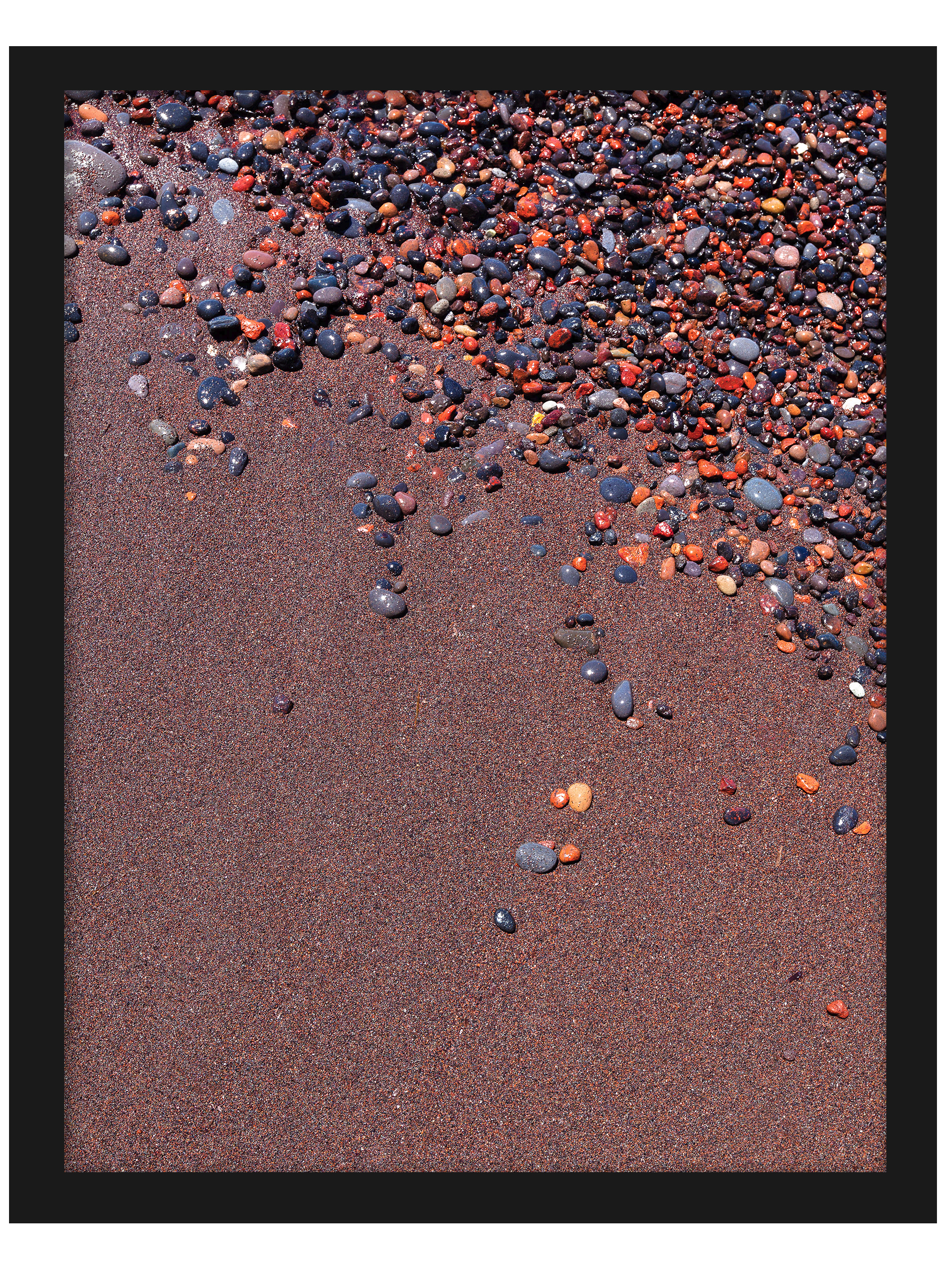 Vibrant red sand and scattered colorful pebbles on a beach in Santorini, Greece.