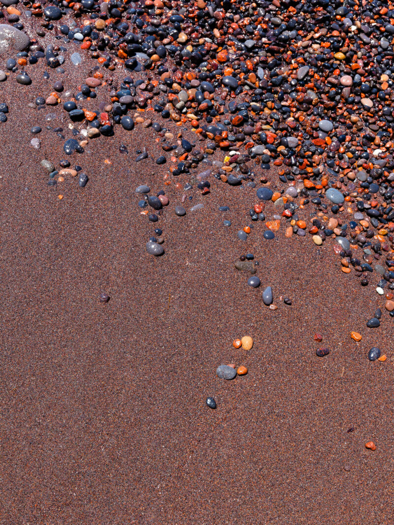 Vibrant red sand and scattered colorful pebbles on a beach in Santorini, Greece.