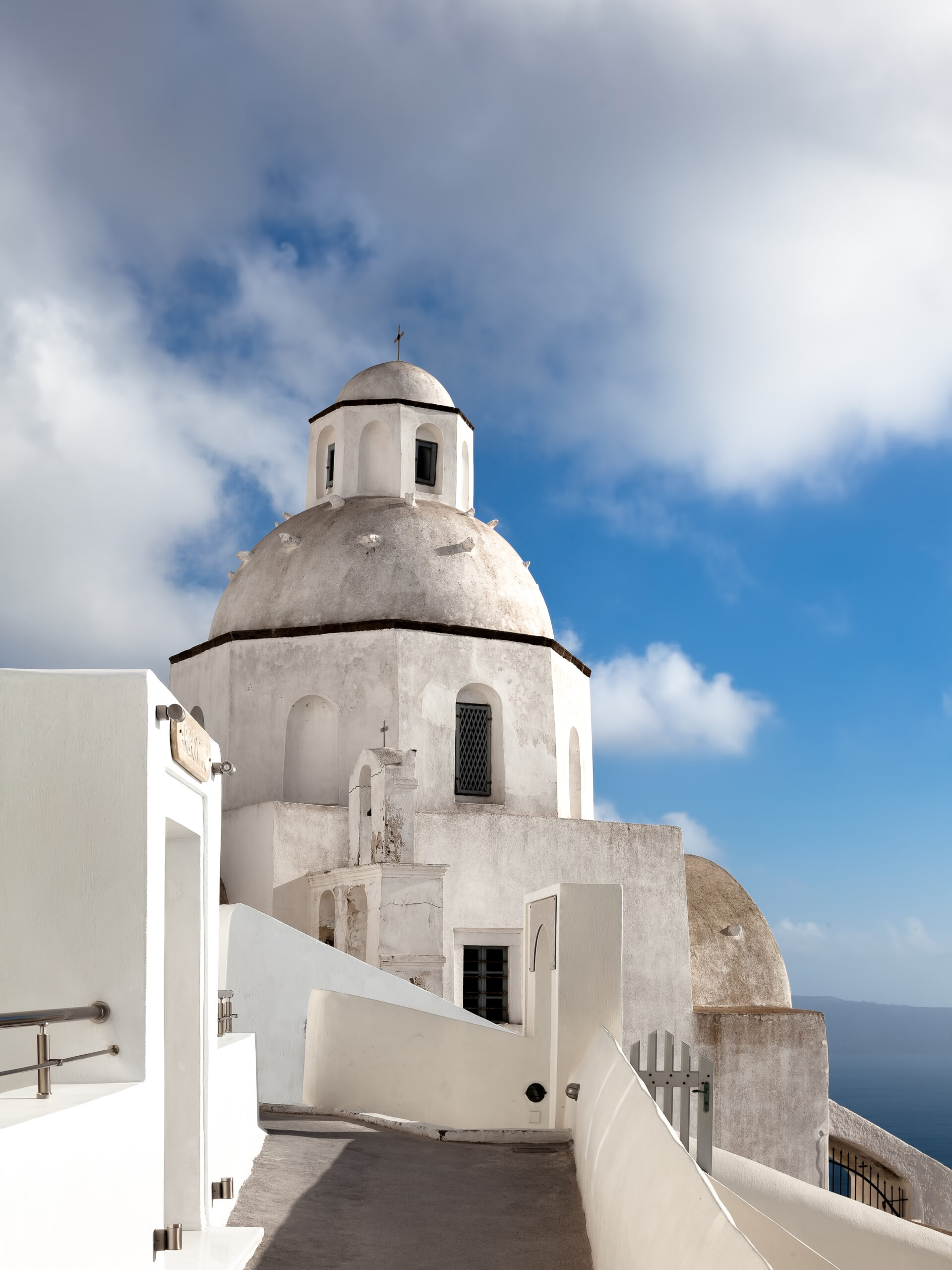 A scenic walkway in Fira, Santorini leading to a white domed church under a bright blue sky.